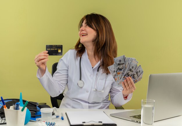 Medico femminile di mezza età gioioso che indossa veste medica e stetoscopio seduto alla scrivania con strumenti medici e computer portatile che tiene soldi e carta di credito guardando lato isolato