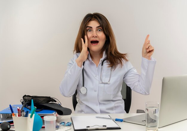 Medico femminile di mezza età impressionato che indossa veste medica e stetoscopio seduto alla scrivania con appunti di strumenti medici e laptop rivolto verso l'alto sussurrando isolato