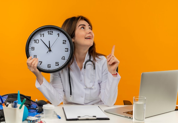 Medico femminile di mezza età impressionato che indossa veste medica e stetoscopio seduto alla scrivania con appunti di strumenti medici e laptop tenendo l'orologio guardando il lato rivolto verso l'alto isolato