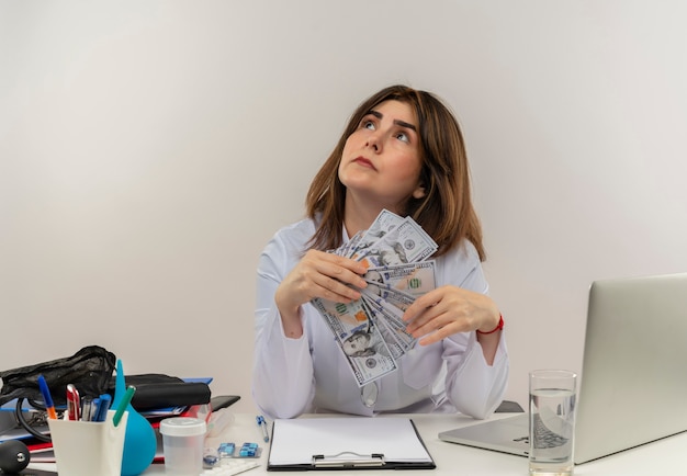 Medico femminile di mezza età premuroso che indossa veste medica e stetoscopio seduto alla scrivania con appunti di strumenti medici e laptop tenendo i soldi guardando in alto isolato