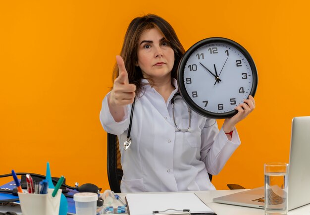 Medico femminile di mezza età sicuro che indossa veste medica e stetoscopio seduto alla scrivania con appunti di strumenti medici e laptop con orologio che indica isolato