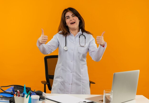 Medico femminile di mezza età soddisfatto che indossa la veste medica con lo stetoscopio che si siede alla scrivania lavora sul computer portatile con strumenti medici i suoi pollici in su sulla parete arancione isolata con lo spazio della copia