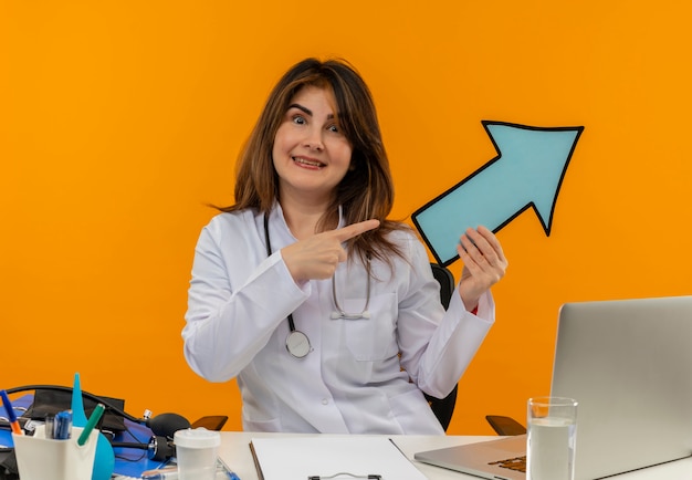 Medico femminile di mezza età sorridente che indossa veste medica e stetoscopio seduto alla scrivania con appunti di strumenti medici e laptop tenendo premuto il segno di freccia che punta a lato isolato