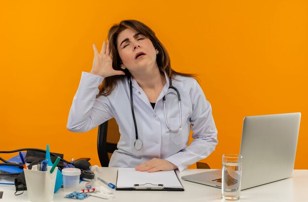 Medico femminile di mezza età stanco che indossa la veste medica con lo stetoscopio che si siede alla scrivania lavora sul computer portatile con strumenti medici mettendo la mano sulla testa su fondo arancione isolato con lo spazio della copia