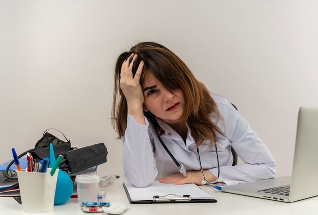 Medico femminile di mezza età stanco che indossa veste medica e stetoscopio seduto alla scrivania con appunti di strumenti medici e laptop mettendo la mano sulla testa cercando isolato