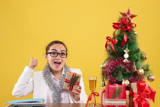 Medico femminile di vista frontale che si siede con i regali di Natale e l'albero su fondo giallo