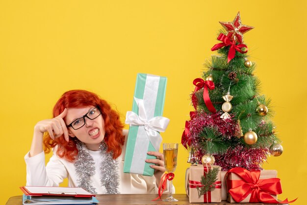 Medico femminile di vista frontale che si siede con i regali di Natale e l'albero su fondo giallo