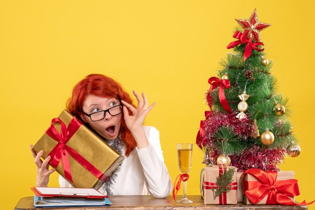 Medico femminile di vista frontale che si siede con i regali di Natale e l'albero su fondo giallo