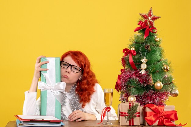 Medico femminile di vista frontale che si siede con i regali di Natale e l'albero su fondo giallo