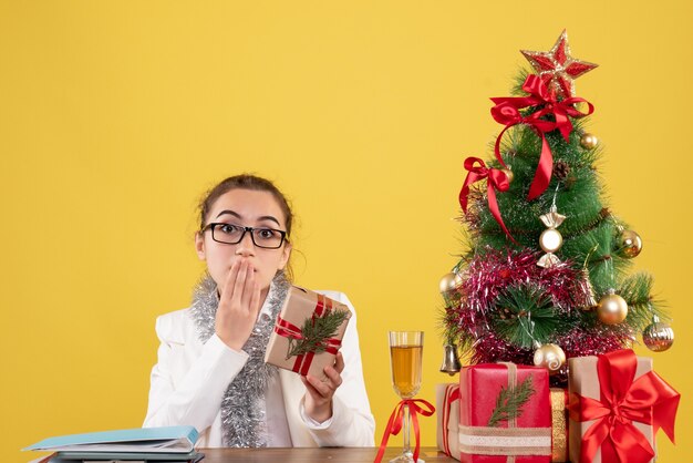 Medico femminile di vista frontale che si siede con i regali di Natale e l'albero su fondo giallo