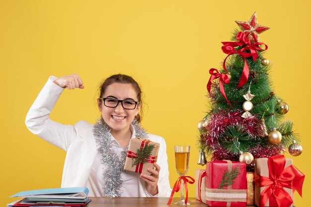 Medico femminile di vista frontale che si siede con i regali di Natale e l'albero su fondo giallo