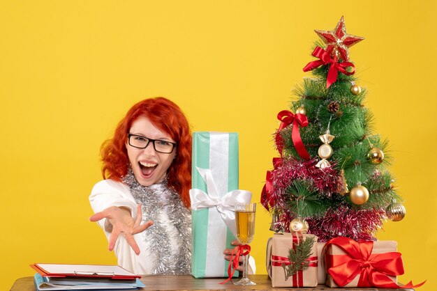 Medico femminile di vista frontale che si siede con i regali di Natale e l'albero su fondo giallo