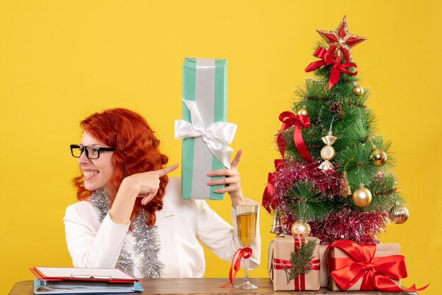 Medico femminile di vista frontale che si siede con i regali di Natale e l'albero sullo scrittorio giallo