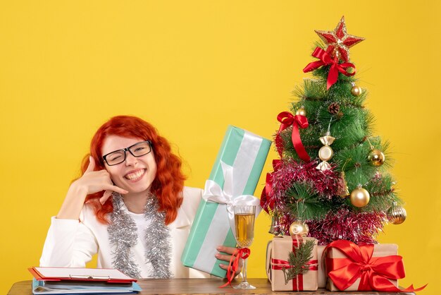 Medico femminile di vista frontale che si siede con i regali di Natale e l'albero sullo scrittorio giallo