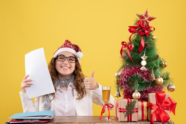 Medico femminile di vista frontale che si siede con l'albero dei regali di Natale e che tiene i documenti su fondo giallo