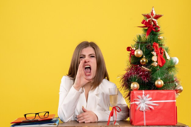 Medico femminile di vista frontale che si siede davanti al suo tavolo che chiama con rabbia su sfondo giallo con albero di Natale e scatole regalo