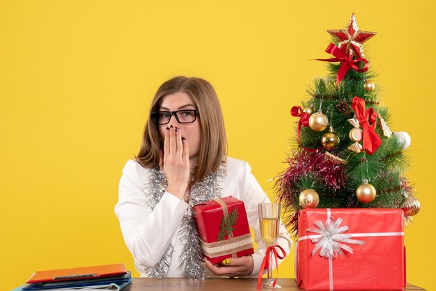 Medico femminile di vista frontale che si siede davanti al suo tavolo che tiene presente su sfondo giallo con albero di Natale e scatole regalo