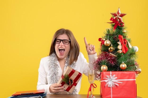 Medico femminile di vista frontale che si siede davanti al suo tavolo che tiene presente su sfondo giallo con albero di Natale e scatole regalo