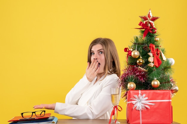 Medico femminile di vista frontale che si siede davanti al suo tavolo su sfondo giallo con albero di Natale e scatole regalo