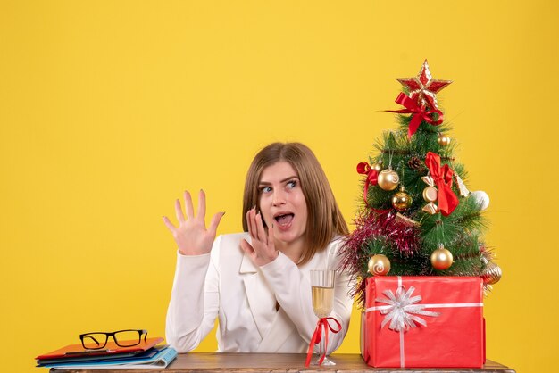 Medico femminile di vista frontale che si siede davanti al tavolo con regali di Natale e albero su sfondo giallo