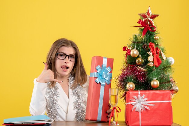 Medico femminile di vista frontale che si siede davanti al tavolo con regali e albero sullo scrittorio giallo con albero di Natale e scatole regalo