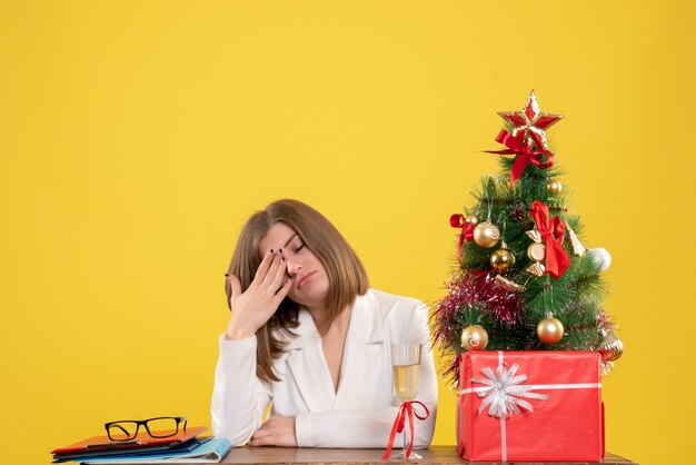 Medico femminile di vista frontale che si siede davanti alla sua tavola sullo scrittorio giallo con i contenitori di regalo e dell'albero di Natale