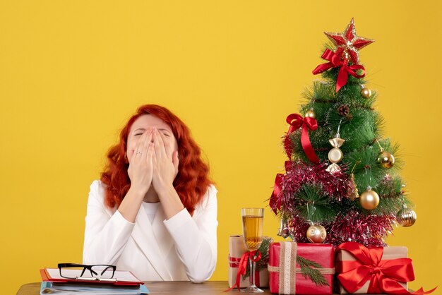 Medico femminile di vista frontale che si siede davanti alla tavola con i regali e l'albero di Natale che sbadiglia su fondo giallo