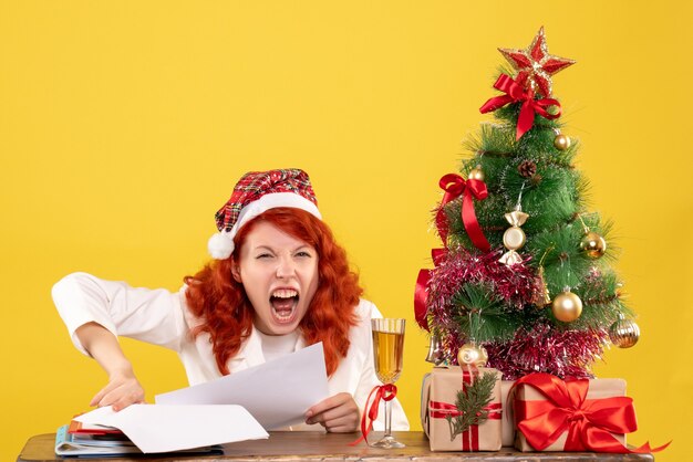 Medico femminile di vista frontale che si siede dietro il tavolo con regali e urlando su sfondo giallo con albero di Natale e scatole regalo