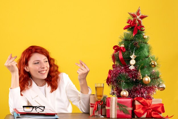 Medico femminile di vista frontale che si siede dietro la sua tavola con i regali di Natale e l'albero su fondo giallo