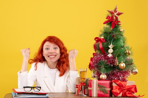 Medico femminile di vista frontale che si siede dietro la tavola con i regali di Natale su colore giallo