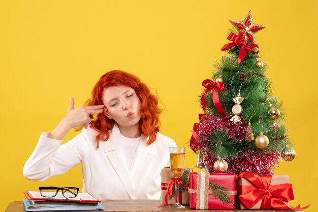 Medico femminile di vista frontale che si siede dietro la tavola con i regali di Natale su fondo giallo