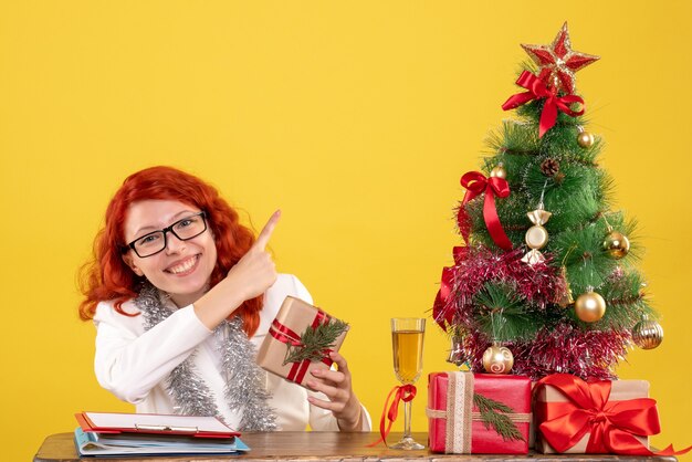 Medico femminile di vista frontale che si siede dietro la tavola con i regali di Natale su fondo giallo