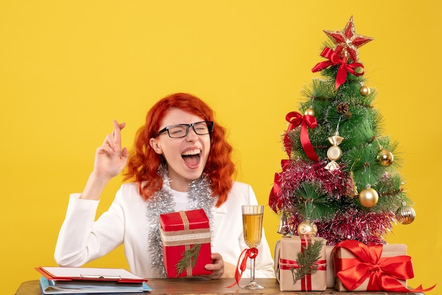 Medico femminile di vista frontale che si siede dietro la tavola con i regali di Natale su fondo giallo