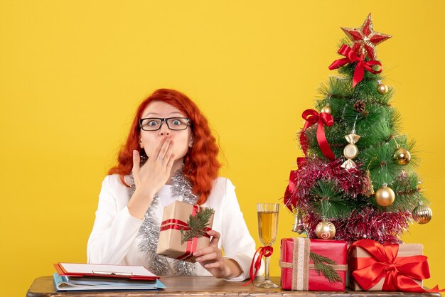 Medico femminile di vista frontale che si siede dietro la tavola con i regali di Natale su fondo giallo