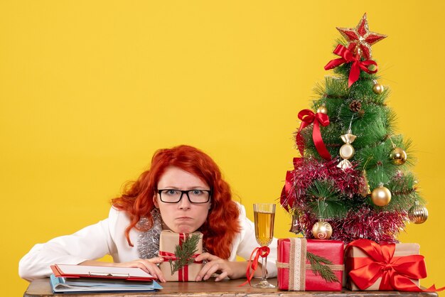 Medico femminile di vista frontale che si siede dietro la tavola con i regali di Natale su fondo giallo