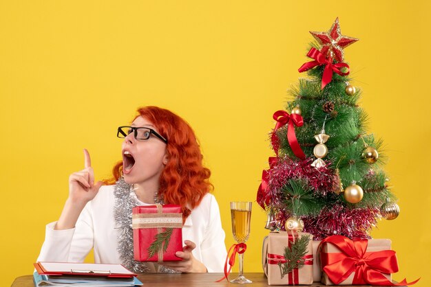 Medico femminile di vista frontale che si siede dietro la tavola con i regali di Natale su fondo giallo