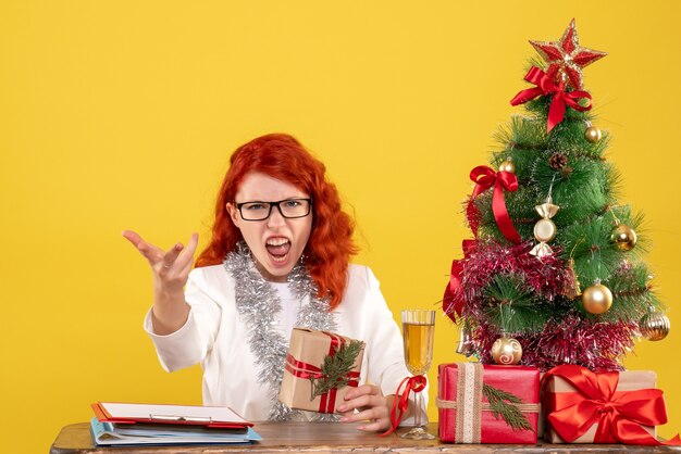 Medico femminile di vista frontale che si siede dietro la tavola con i regali di Natale su fondo giallo