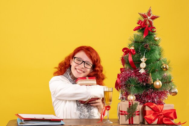 Medico femminile di vista frontale che si siede dietro la tavola con i regali di Natale sullo scrittorio giallo con i contenitori di regalo e dell'albero di Natale