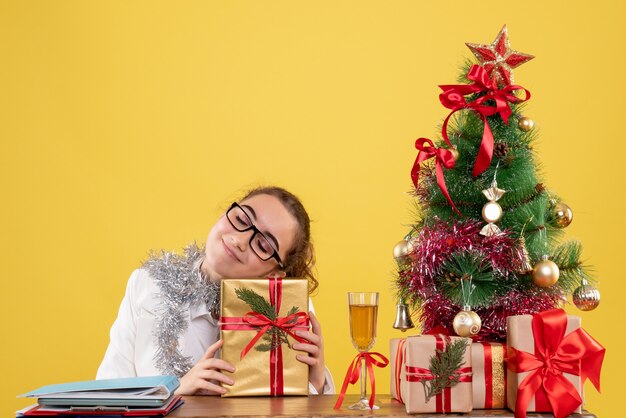 Medico femminile di vista frontale che si siede intorno ai regali di Natale e all'albero con la faccia felice su fondo giallo