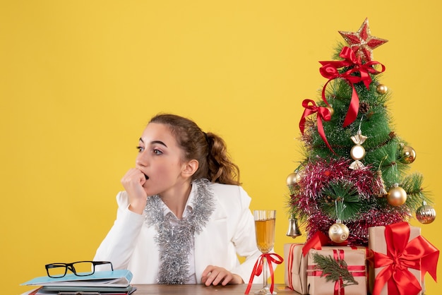Medico femminile di vista frontale che si siede intorno ai regali di Natale e all'albero su fondo giallo