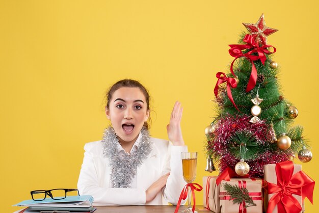 Medico femminile di vista frontale che si siede intorno ai regali di Natale e all'albero su fondo giallo