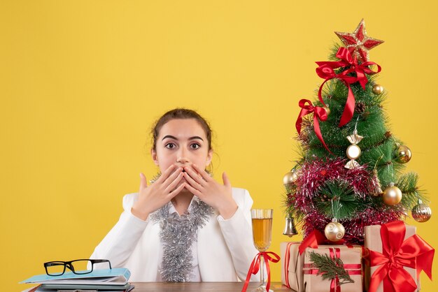 Medico femminile di vista frontale che si siede intorno ai regali di Natale e all'albero su fondo giallo