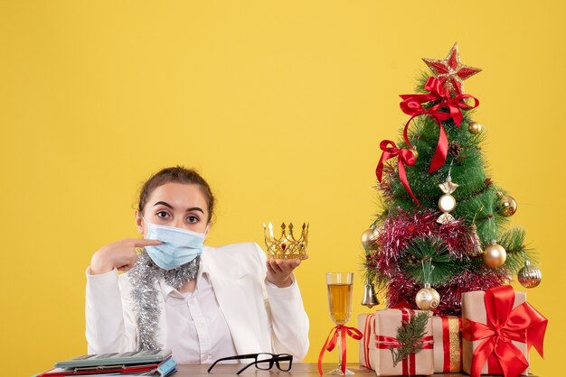 Medico femminile di vista frontale che si siede nella corona sterile della tenuta della maschera su fondo giallo con l'albero di Natale e le confezioni regalo