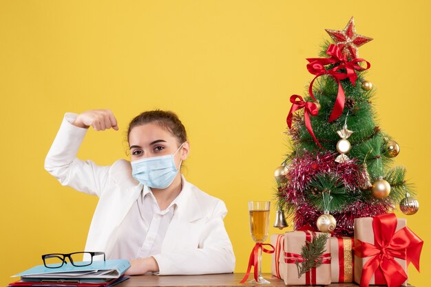 Medico femminile di vista frontale che si siede nella maschera sterile che flette su fondo giallo con l'albero di Natale e le confezioni regalo