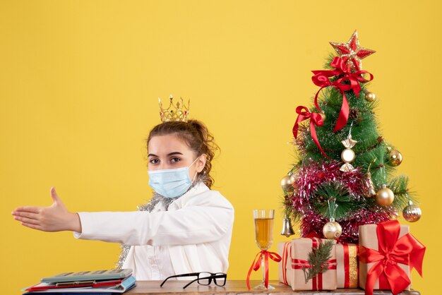 Medico femminile di vista frontale che si siede nella maschera sterile su fondo giallo con l'albero di Natale e confezioni regalo