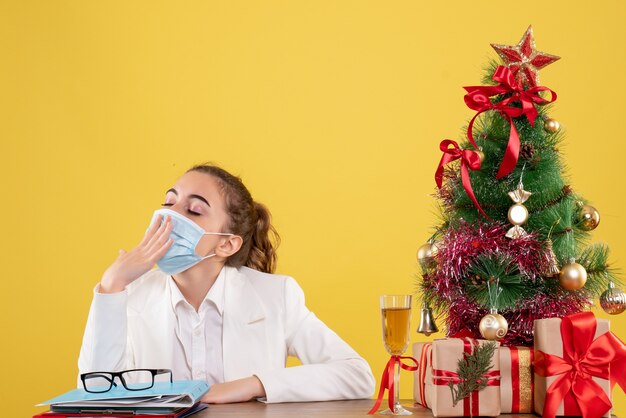 Medico femminile di vista frontale che si siede nella mascherina protettiva che sbadiglia su priorità bassa gialla con i contenitori di regalo e dell'albero di Natale
