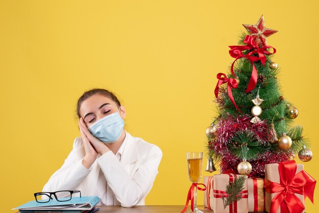 Medico femminile di vista frontale che si siede nella mascherina protettiva che si sente stanco su fondo giallo con l'albero di Natale e le confezioni regalo