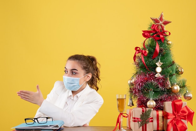 Medico femminile di vista frontale che si siede nella mascherina protettiva su priorità bassa gialla con l'albero di Natale e confezioni regalo