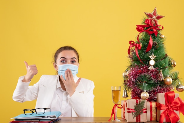 Medico femminile di vista frontale che si siede nella mascherina protettiva su priorità bassa gialla con l'albero di Natale e confezioni regalo