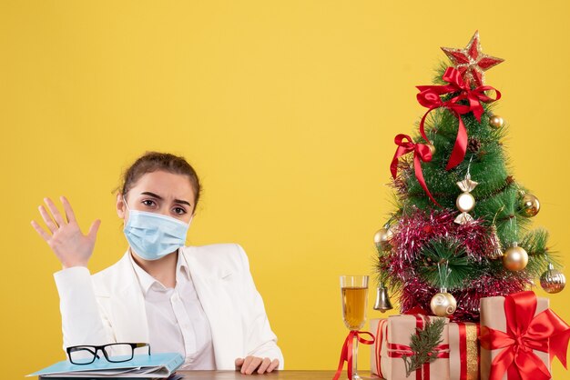 Medico femminile di vista frontale che si siede nella mascherina protettiva su priorità bassa gialla con l'albero di Natale e confezioni regalo